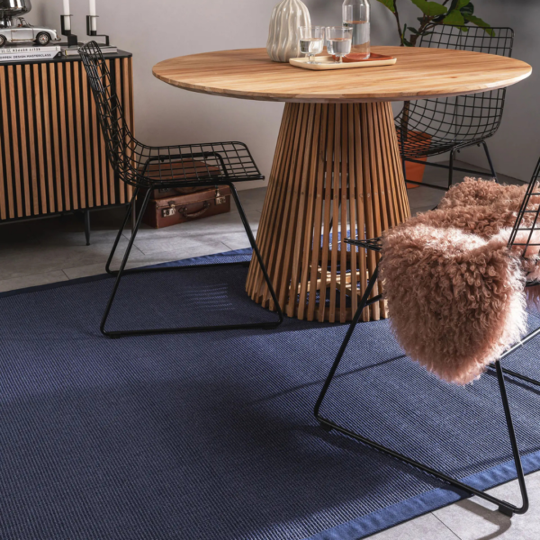 Ein Bild aus dem Taiga Store von taiga.green. Moderner Essbereich mit rundem Holztisch mit Lattenrost, Stühlen mit Drahtgestell - einer davon mit einer rosa Kunstpelzdecke drapiert - auf einem blauen Teppich, einem gestreiften Sideboard und einem Plattenspieler im Hintergrund. Jetzt bestellen im Eco Shop von www.taiga.green. Hier nachhaltig einkaufen mit dem Taiga Eco Rating